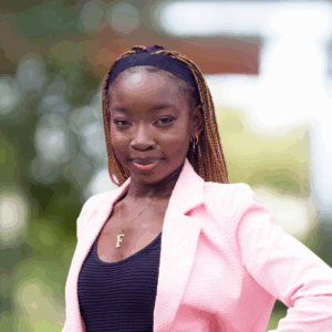 A woman with braided hair wearing a pink blazer over a black top and black pants stands outdoors, smiling confidently with her hand on her hip. The background is blurred greenery.